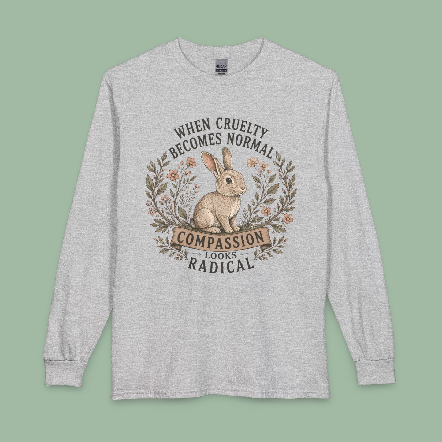 A light heather gray long-sleeve t-shirt with a graphic print of a rabbit inside a wreath and the text 'WHEN CRUELTY BECOMES NORMAL COMPASSION LOOKS RADICAL'. The shirt is displayed flat against a solid sage green background.