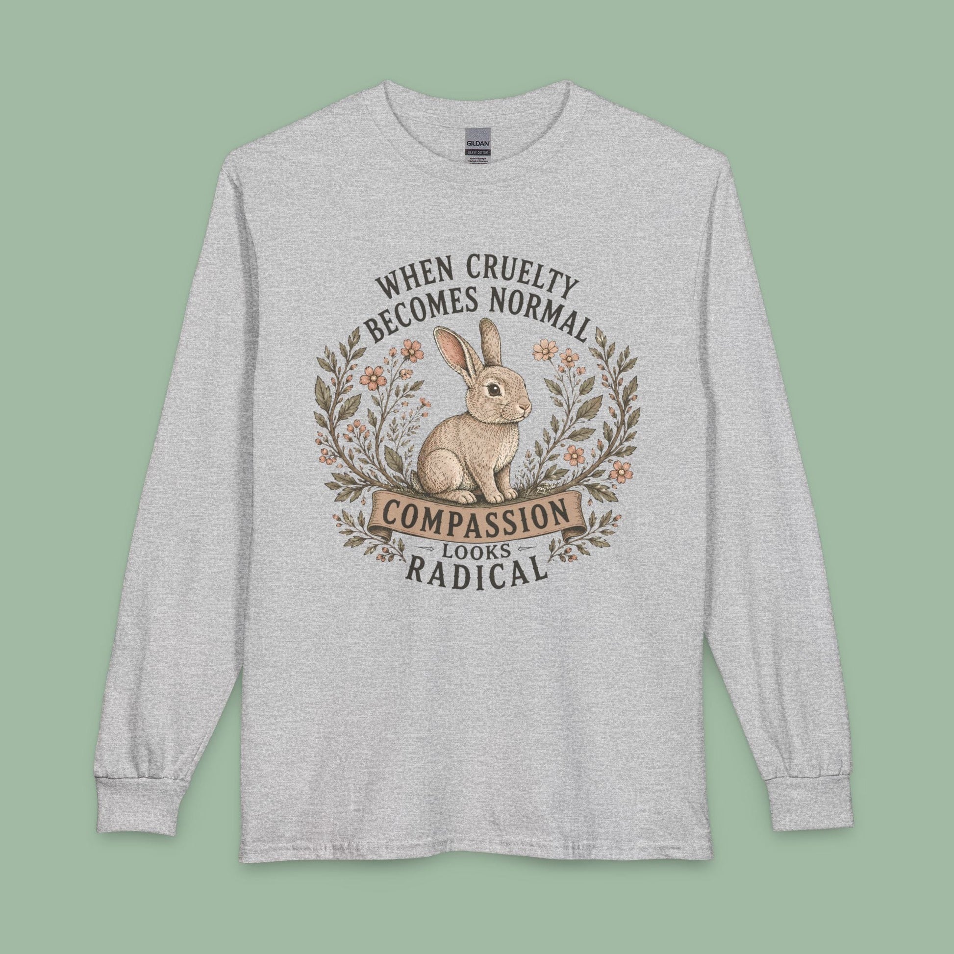 A light heather gray long-sleeve t-shirt with a graphic print of a rabbit inside a wreath and the text 'WHEN CRUELTY BECOMES NORMAL COMPASSION LOOKS RADICAL'. The shirt is displayed flat against a solid sage green background.