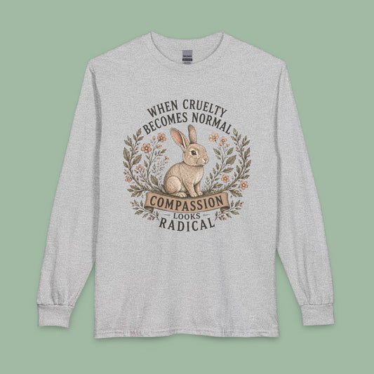 A light heather gray long-sleeve t-shirt with a graphic print of a rabbit inside a wreath and the text 'WHEN CRUELTY BECOMES NORMAL COMPASSION LOOKS RADICAL'. The shirt is displayed flat against a solid sage green background.