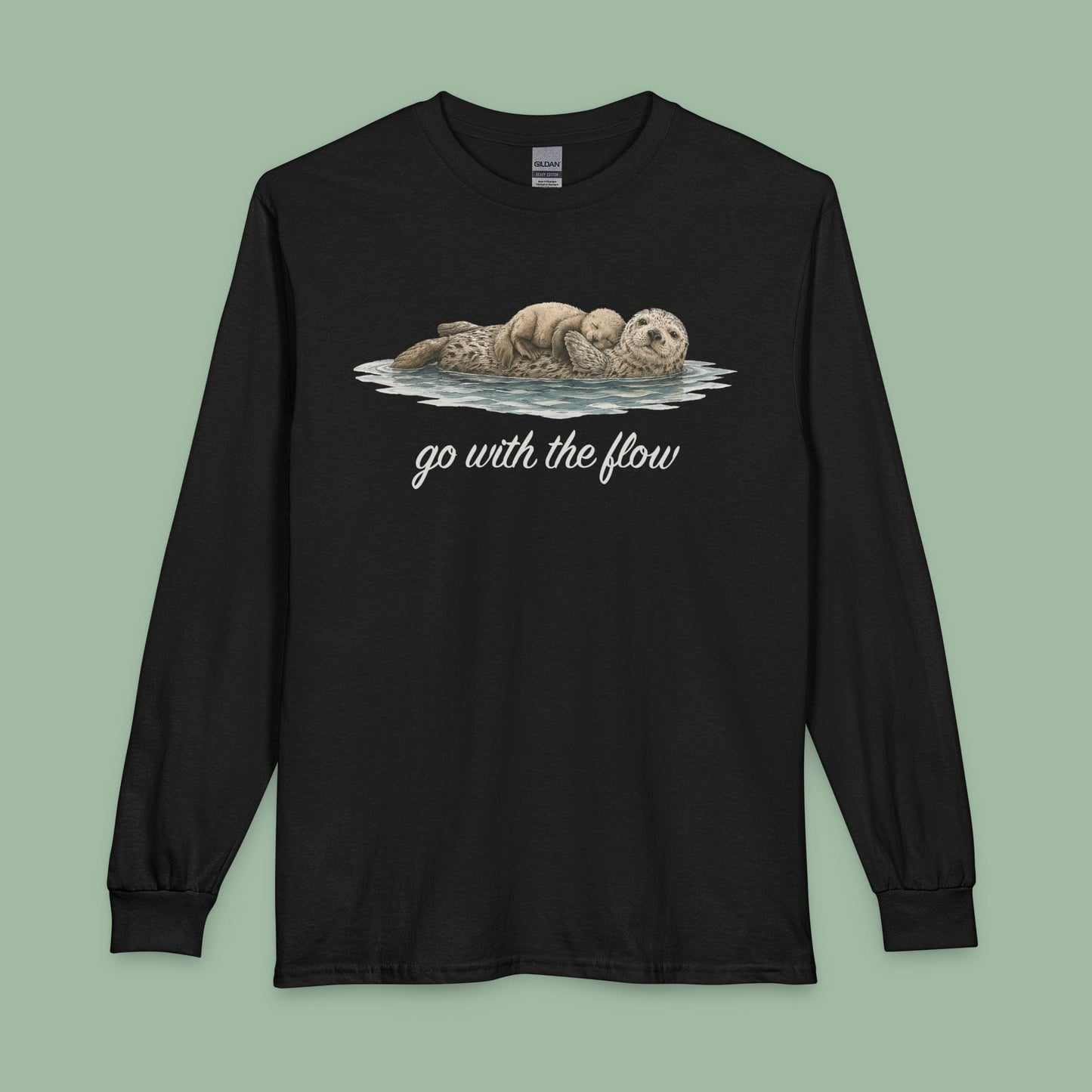 A black long-sleeve crewneck t-shirt featuring a graphic of two sea otters floating on their backs with the text 'go with the flow' printed below in white script, displayed against a solid sage green background.
