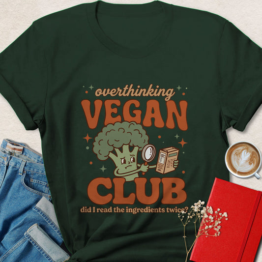 Green t-shirt with 'overthinking VEGAN CLUB' design featuring a broccoli character, placed on a surface with jeans, a coffee cup, and a book.