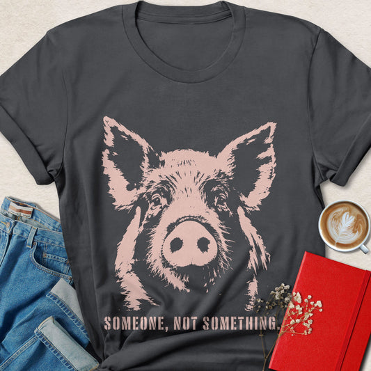 A dark charcoal gray graphic t-shirt featuring a stylized pink pig illustration and the text 'SOMEONE, NOT SOMETHING.' in pink.