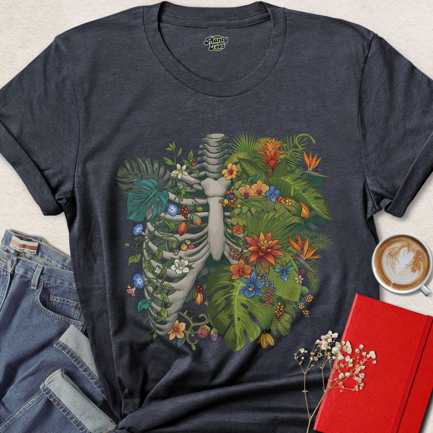Heather Navy blue t-shirt with a floral and skeleton design, placed on a surface with jeans, a coffee cup, and a book.