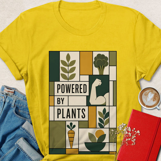 A bright yellow graphic T-shirt laid flat on a light textured surface, styled with jeans, a cup of latte, and a red notebook. The shirt features a large rectangular retro-style print reading 'POWERED BY PLANTS' with illustrated leaves, broccoli, a flexed bicep, a carrot, and other plant motifs.