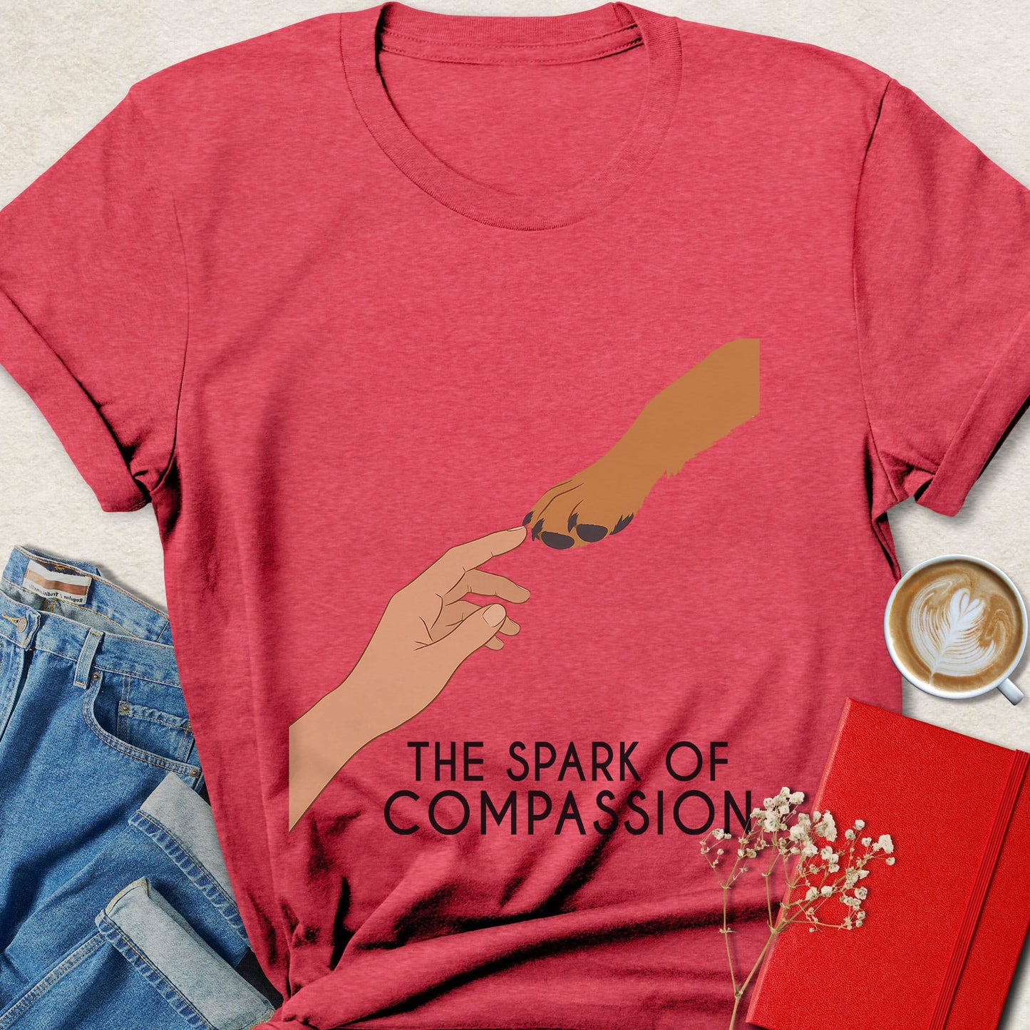A heather red graphic t-shirt featuring an illustration of two hands reaching toward each other with the text 'THE SPARK OF COMPASSION' underneath. The shirt is styled in a flat-lay arrangement with blue jeans, a cup of coffee, a red notebook, and small white flowers on a light beige textured background.
