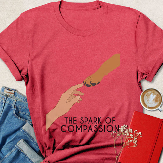 A heather red graphic t-shirt featuring an illustration of two hands reaching toward each other with the text 'THE SPARK OF COMPASSION' underneath. The shirt is styled in a flat-lay arrangement with blue jeans, a cup of coffee, a red notebook, and small white flowers on a light beige textured background.