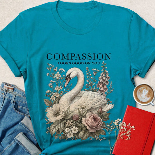 A turquoise short-sleeve graphic T-shirt laid flat on a light neutral surface, styled with jeans, a red notebook, and a cup of latte. The shirt has a large front print of a white swan surrounded by flowers and the text 'COMPASSION LOOKS GOOD ON YOU'.