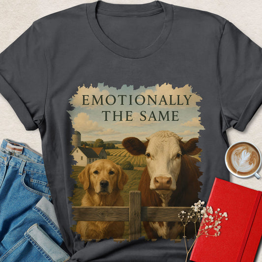 A dark gray graphic t-shirt featuring a printed design of a golden retriever and a brown and white cow in front of a barn, with the text 'EMOTIONALLY THE SAME' above them. The shirt is styled in a flat-lay arrangement with blue jeans, a cup of coffee, a red notebook, and small white flowers on a light textured background.
