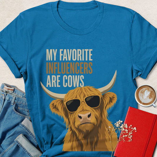 A bright blue graphic T-shirt laid flat with jeans, a coffee cup, and a red notebook around it. The shirt has a large illustration of a brown highland cow wearing sunglasses and the text 'MY FAVORITE INFLUENCERS ARE COWS' in white and tan.