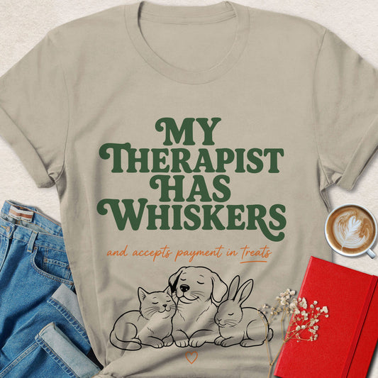 A beige graphic T-shirt laid flat with blue jeans, a cup of latte, a red notebook, and small flowers around it. The shirt reads “MY THERAPIST HAS WHISKERS and accepts payment in treats” above a line drawing of a dog, cat, and rabbit.