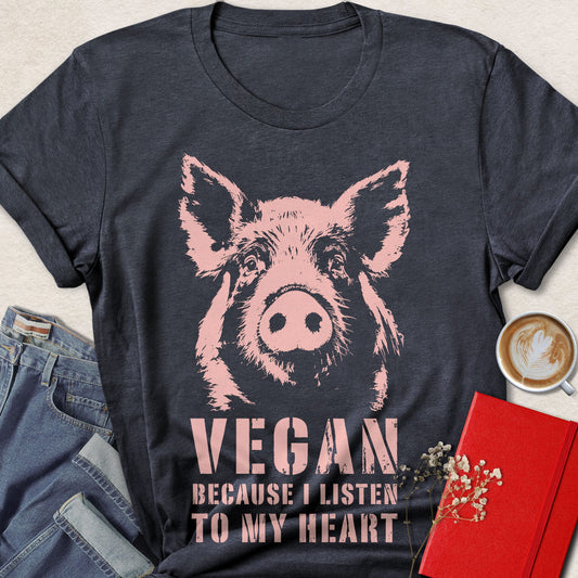 A heather navy graphic t-shirt featuring a pink pig illustration and the text 'VEGAN BECAUSE I LISTEN TO MY HEART'. The t-shirt is styled in a flat-lay arrangement with blue jeans, a cup of coffee, and a red notebook on a light beige textured background.