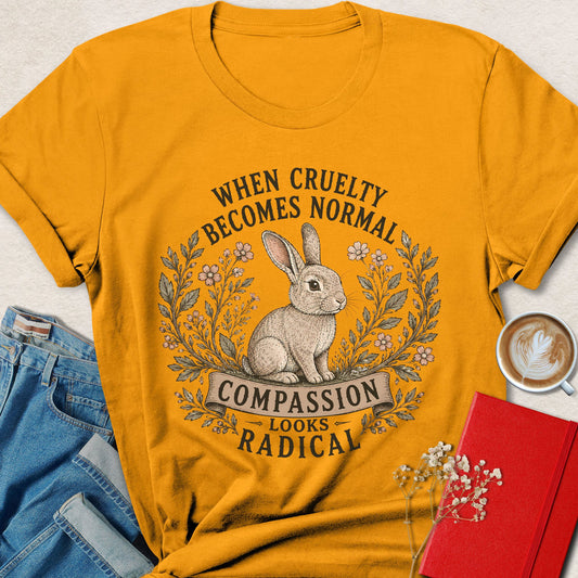 A gold colored graphic t-shirt featuring a rabbit illustration and the text 'WHEN CRUELTY BECOMES NORMAL COMPASSION LOOKS RADICAL'. The shirt is styled in a flat-lay arrangement with blue jeans, a red notebook, and a cup of coffee on a light beige background.